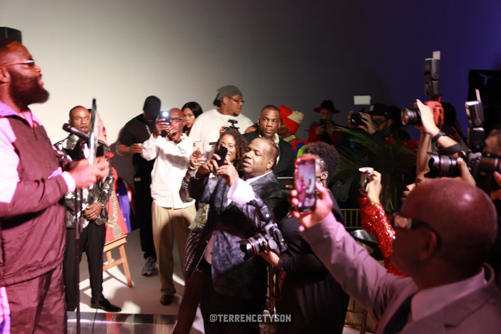 Rick Ross on stage with award in his hand with people taking pictures of him